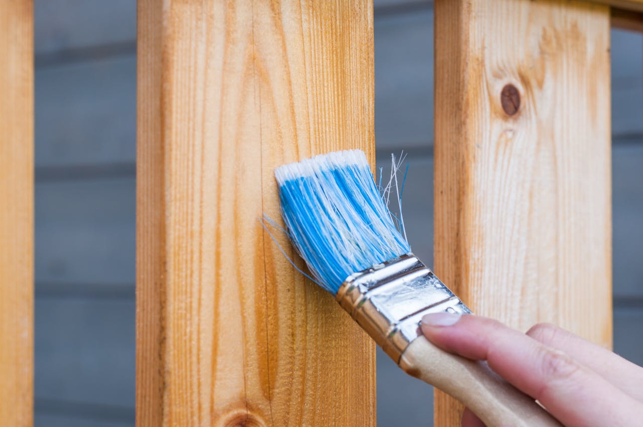 heros-img A hand painting a wooden fence plank with a blue paintbrush outdoors. Perfect for DIY and renovation themes.
